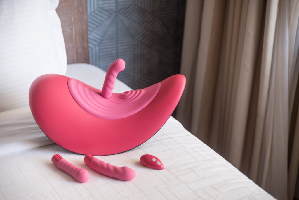 The VVD Rider Desire machine sits out on the edge of a bed. One of its attachments is in it, but the other two attachments and the remote sit next to it. For my VVD Rider Desire review.