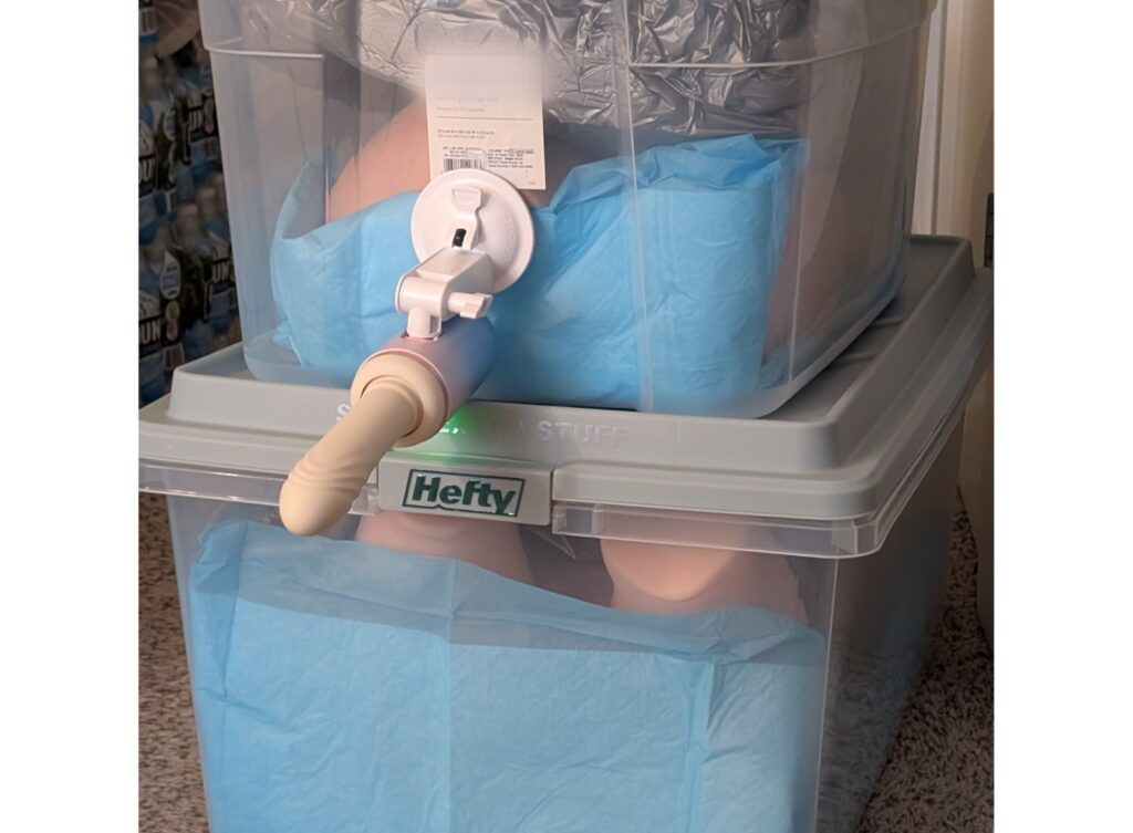 Lovense Spinel sex machine suction cupped to the side of a plastic tote that's stacked on top of another tote. The totes look like they have sex dolls inside. 