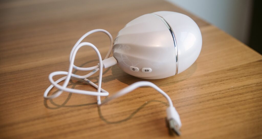 The Cloud Kiss sits on a table with the charging cable plugged into its base. The opposite side of the charging cable has a USB end.