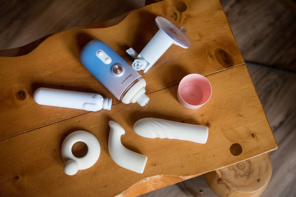 Everything that comes with the Lovense Spinel is laid out on a table. It has a hand holding attachment and a suction cup one. It also has a clitoral bump for pleasure alongside a g-spot dildo and straight dildo. The lid is removed from the machine and sitting nearby. For my Lovense Spinel review.