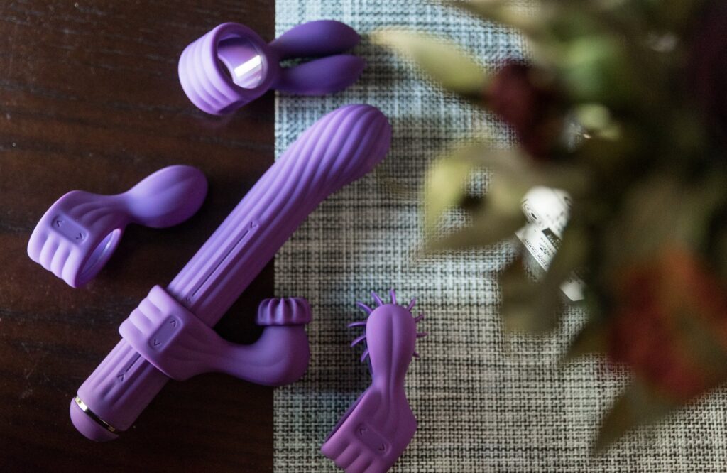 All of the pieces of the 4 attachment rabbit vibrator laid out on a table. There's a green plant in the foreground of the image. For my AVIDLOVE PLAY Four-Attachment Rabbit Vibrator review.