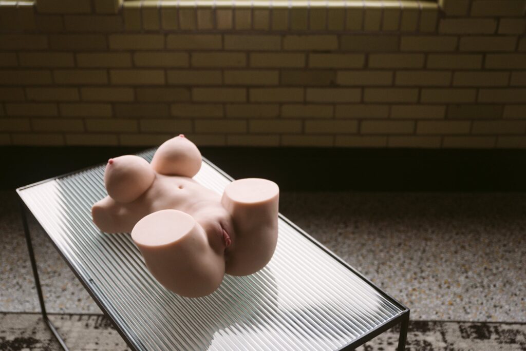 The sex doll sits out on a glass table. The angle has the vulva closest to the camera with a smooth appearance. For my Salorein Sabrina review.