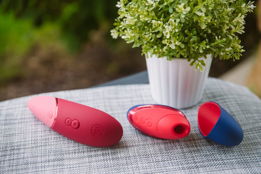 Both of the toys sit outdoors on a gray placement with green bushes in the background. The toys are extremely bright, colorful, and welcoming. For my Womanizer Next Liberty vs Womanizer Next comparison