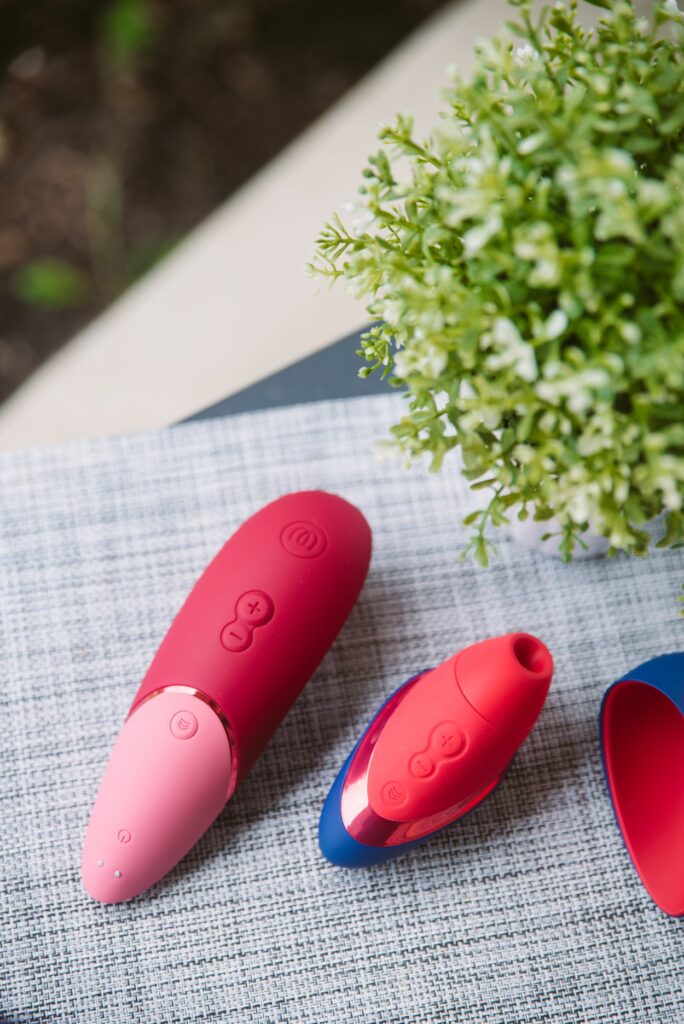 Both of the toys sit outdoors on a gray placement with green bushes in the background. The toys are extremely bright, colorful, and welcoming. For my Womanizer Next Liberty vs Womanizer Next comparison