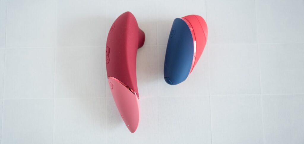 The two sex toys are laid on their sides next to one another. The Womanizer Next is noticeably taller in height but slightly thinner in width.