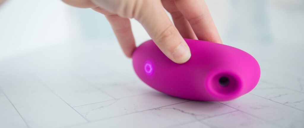 My hand holds the toy on its side, showcasing the air suction hole in addition to the LED light that illuminates the single control button on the base. For my AVIDLOVE PLAY Ergonomic Max Suction Clitoral Vibrator review.