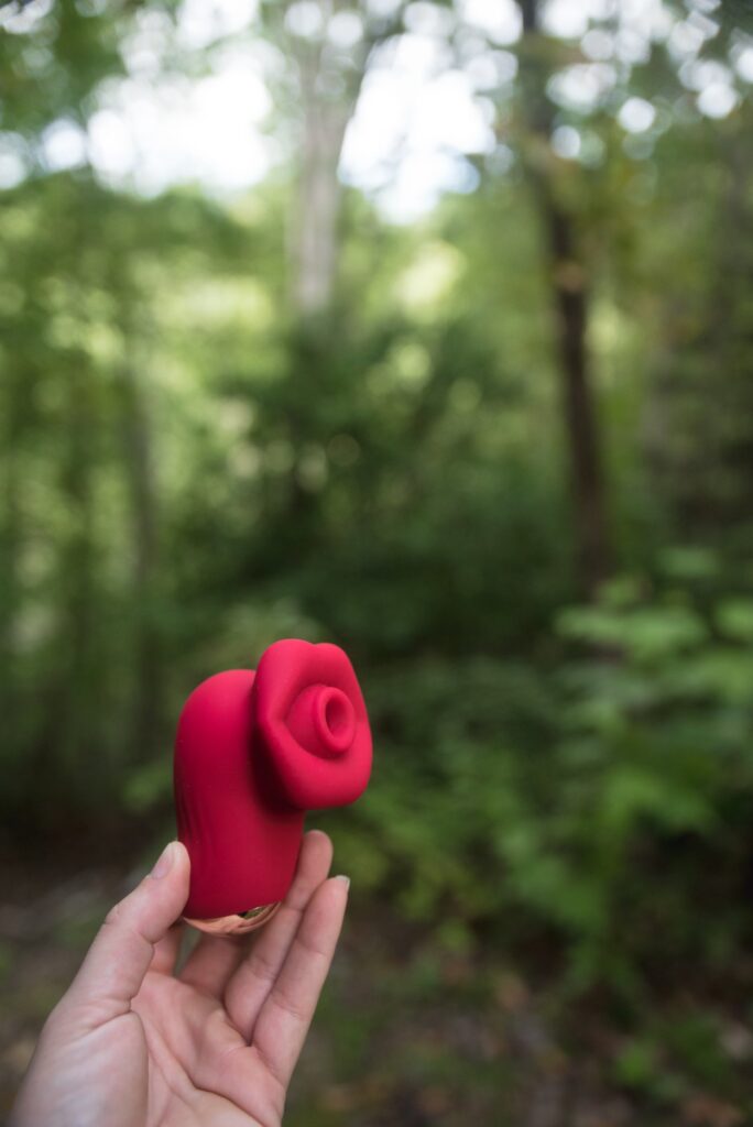 My hand holds the vibrator with a duck-bill like grip at the base. In the background, there's a forest. Everything is evenly lit, and the toy looks extremely high-quality and welcoming. For my AVIDLOVE PLAY Kiss Air Pulse Vibrator Review.